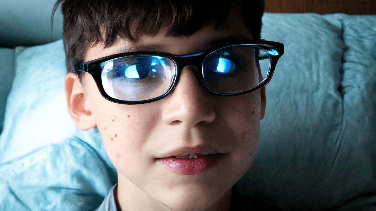 “A close-up of a teenage boy with braces and soft freckles, his face lit by the glow of his phone, reflections shimmering across his glasses, the bedroom around him fading into cool, grainy shadows, youthful, quiet, contemplative.”